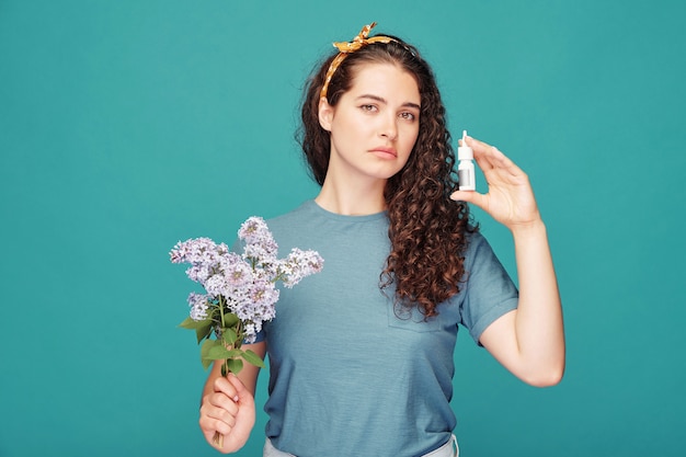 Giovane femmina con ramo di lilla ti guarda e mostra un piccolo contenitore di plastica con un efficace spray nasale antiallergico in isolamento