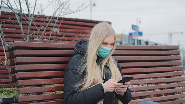 Giovane femmina in una mascherina medica che si siede sul banco e usando il telefono