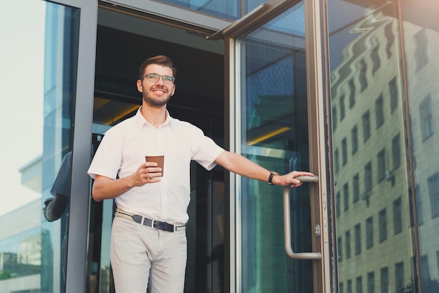 Giovane fiducioso con il telefono cellulare che apre la porta e esce dal moderno edificio per uffici