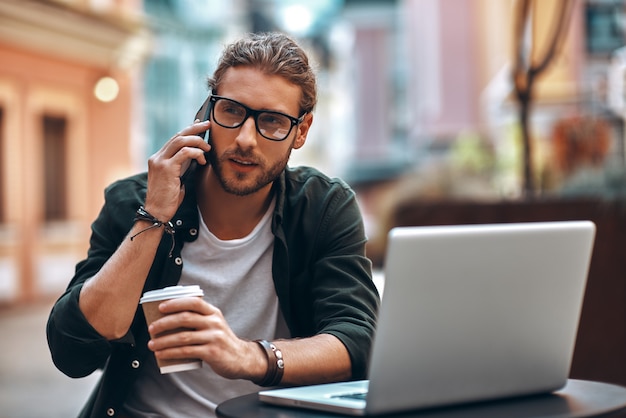 Giovane fiducioso in abbigliamento casual che parla allo smartphone e usa il laptop mentre si lavora al bar