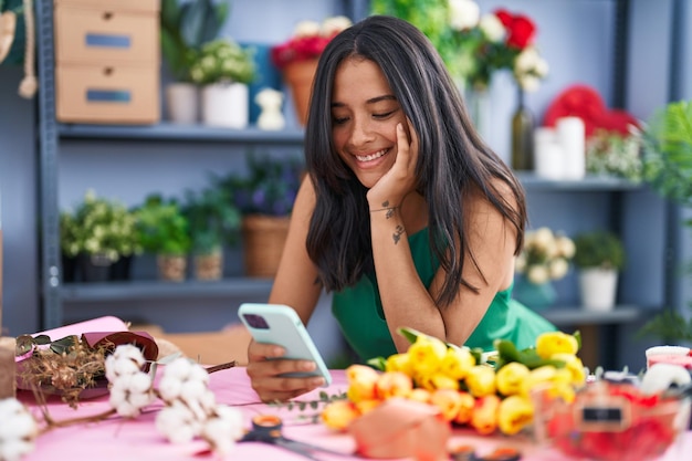 Giovane fiorista donna ispanica sorridente fiducioso utilizzando lo smartphone al negozio di fiori