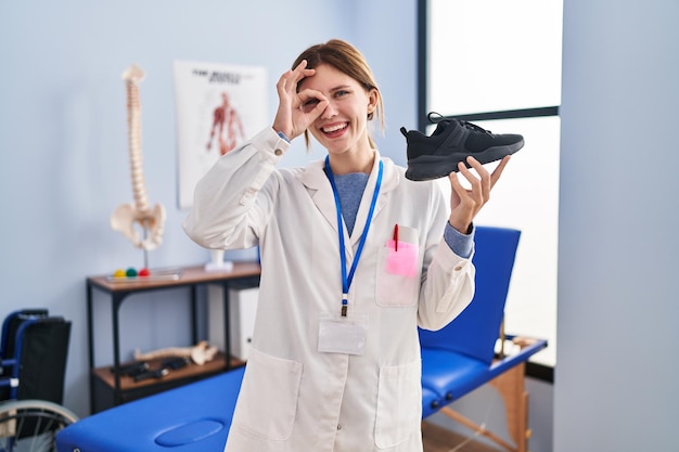 Giovane fisioterapista donna che tiene scarpe da ginnastica sorridendo felice facendo segno ok con la mano sull'occhio guardando attraverso le dita