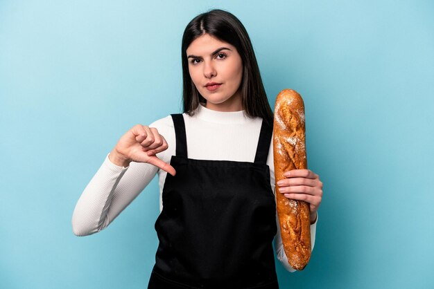 Giovane fornaio caucasico che tiene una pagnotta di pane isolata su sfondo blu che mostra un gesto di antipatia pollice verso il concetto di disaccordo