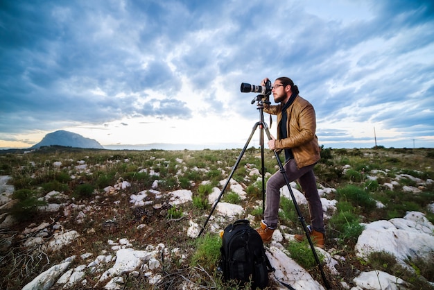 Giovane fotografo attraente che fa le foto con la macchina fotografica sul treppiede al tramonto