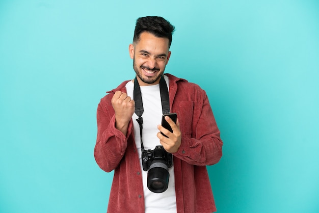 Giovane fotografo caucasico uomo isolato su sfondo blu con telefono in posizione di vittoria