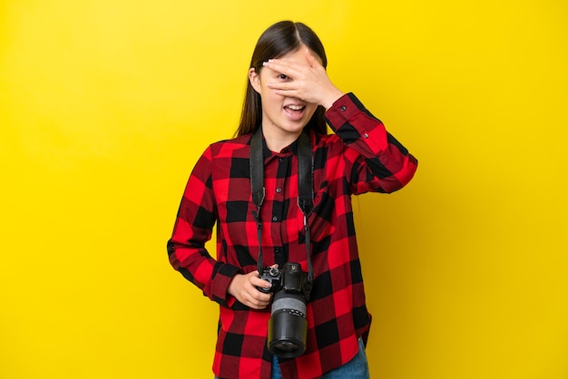 Giovane fotografo donna cinese isolata su sfondo giallo che copre gli occhi con le mani e sorridente