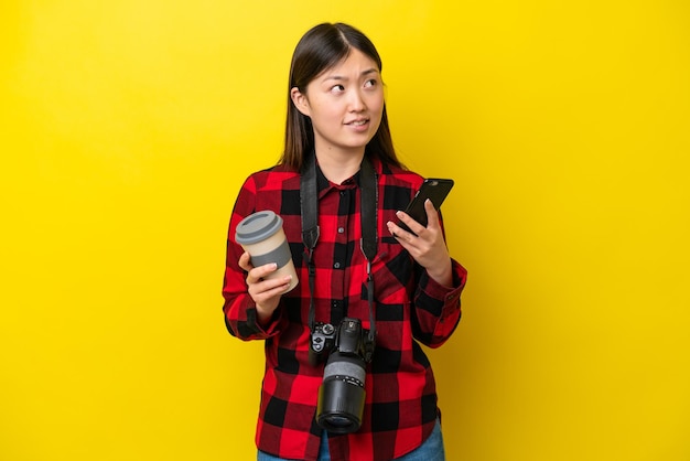 Giovane fotografo donna cinese isolata su sfondo giallo che tiene il caffè da portare via e un cellulare mentre pensa a qualcosa