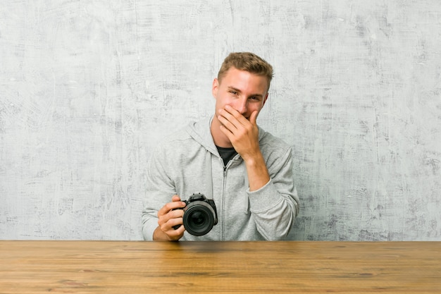 Giovane fotografo in possesso di una macchina fotografica su un tavolo ridendo di qualcosa, coprendo la bocca con le mani.