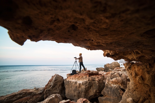 Giovane fotografo maschio che sta in una bocca della caverna della roccia con il treppiede e che fa le foto