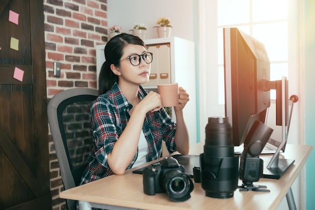 giovane fotografo piuttosto femminile della società per azioni che tiene una tazza di caffè caldo seduto nella scrivania di lavoro e guardando il computer pensando al design dell'immagine.