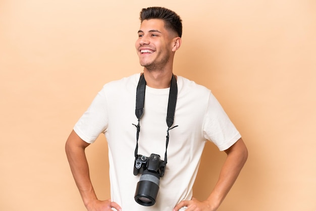 Giovane fotografo uomo caucasico isolato su sfondo beige in posa con le braccia all'anca e sorridente