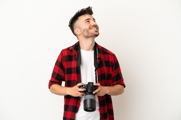 Giovane fotografo uomo caucasico isolato su sfondo bianco guardando in alto mentre sorride