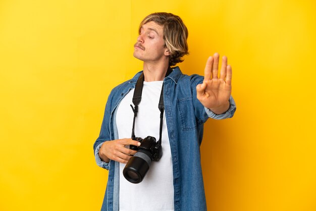 Giovane fotografo uomo isolato sulla parete gialla che fa gesto di arresto e deluso