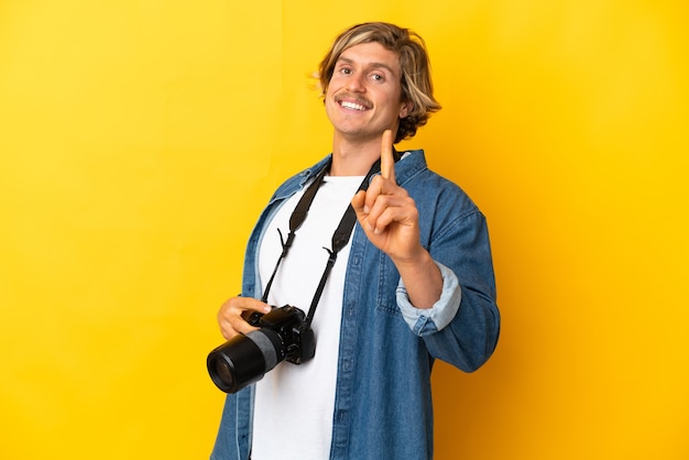 Giovane fotografo uomo isolato sulla parete gialla che mostra e alza un dito