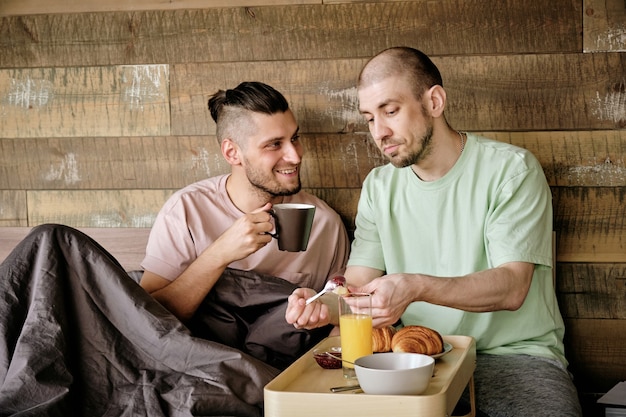 Giovane gay che mette marmellata sul croissant quando il suo ragazzo beve una tazza di caffè e scherza