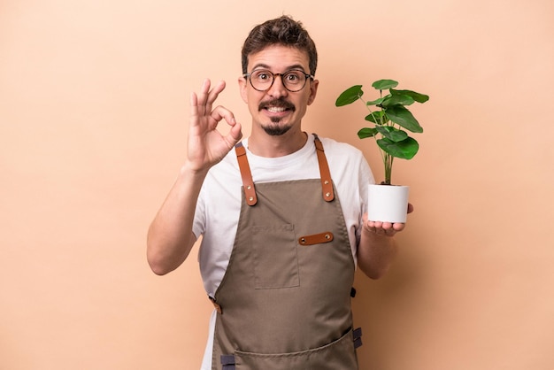 Giovane giardiniere caucasico che tiene una pianta isolata su sfondo beige allegro e fiducioso che mostra un gesto ok