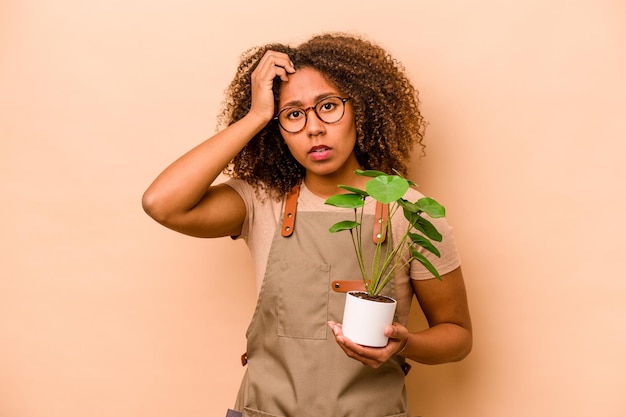 Giovane giardiniere donna afroamericana che tiene pianta isolata su sfondo beige essendo scioccata ha ricordato un incontro importante