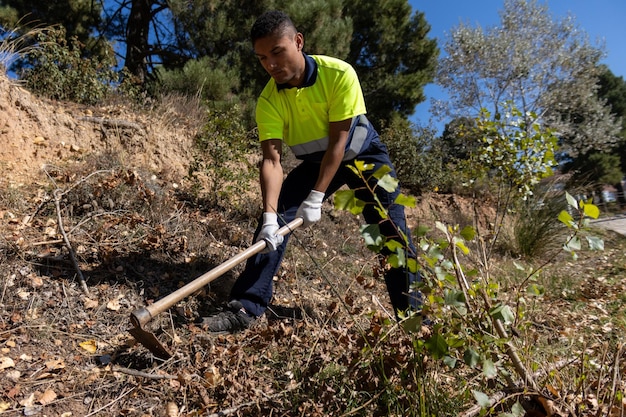 Giovane giardiniere maschio latino in abiti da lavoro riflettenti che scavano e sgombrano la terra per piantare