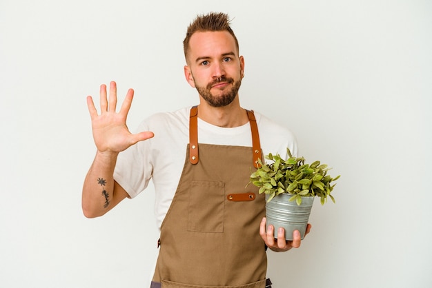 Giovane giardiniere tatuato uomo caucasico che tiene una pianta isolata sul muro bianco sorridente allegro che mostra numero cinque con le dita.