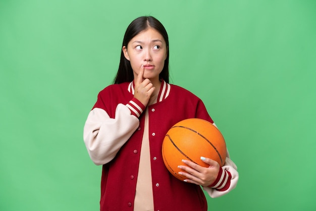 Giovane giocatore di basket donna asiatica su sfondo isolato avendo dubbi e pensando