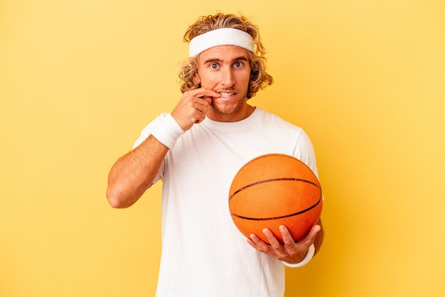 Giovane giocatore di basket uomo caucasico isolato su sfondo giallo che si morde le unghie, nervoso e molto ansioso.