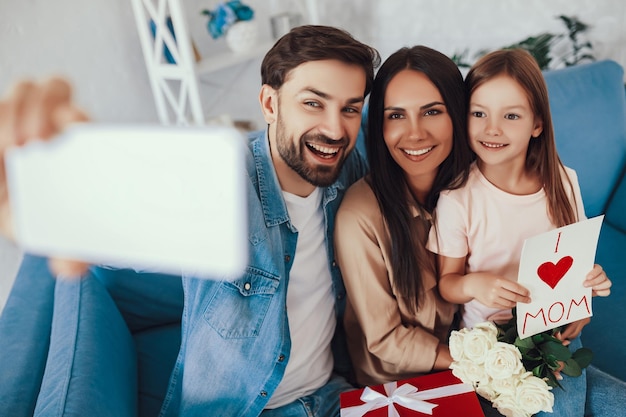 Giovane gioioso che tiene uno smartphone mentre si fa un selfie con la sua bella moglie e la figlia carina