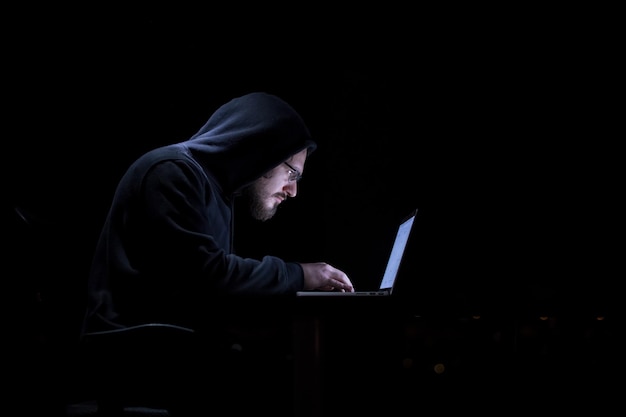 Giovane hacker di talento che utilizza un computer portatile mentre lavora in un ufficio buio con le grandi luci della città sullo sfondo di notte
