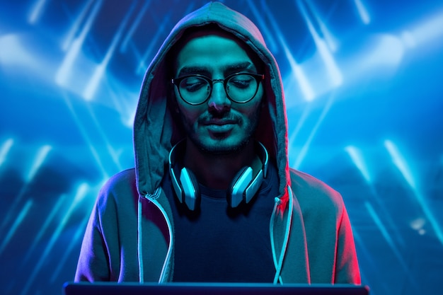 Giovane hacker in felpa con cappuccio e occhiali che irrompono nel sistema informatico su sfondo blu illuminato con linee geometriche