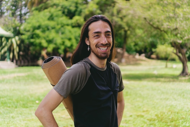 Giovane hippie ispanico latino con i capelli lunghi che sorride mentre guarda la telecamera portando il suo tappetino da yoga