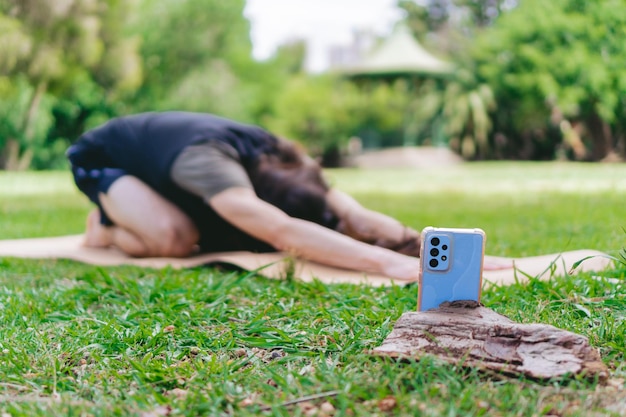 Giovane hippie latino che si filma con il suo smartphone mentre pratica yoga su un tappetino yoga all'aperto