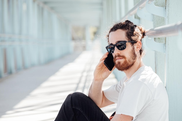 Giovane hipster all'aeroporto o alla stazione degli autobus in attesa mentre chiama qualcuno con il telefono, i bagagli, le borse e la valigia. Giovane viaggiatore hipster capelli lunghi con occhiali da sole, copia spazio, giornata di sole