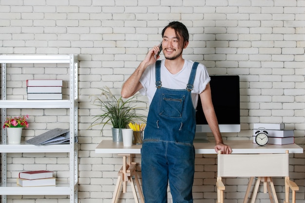 Giovane hipster barba uomo asiatico in piedi può fare una chiamata su smartphone con felice nel suo nuovo ufficio di avvio minimale moderno.