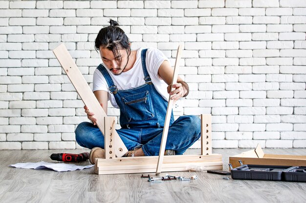 Giovane hipster che lavora come tuttofare, assemblando un tavolo in legno con attrezzature e manuale, concetto per la casa fai da te e self service con muro di mattoni bianchi sullo sfondo.