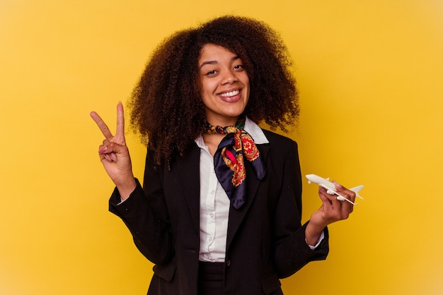 Giovane hostess afroamericana in possesso di un piccolo aereo isolato su sfondo giallo che mostra il numero due con le dita.