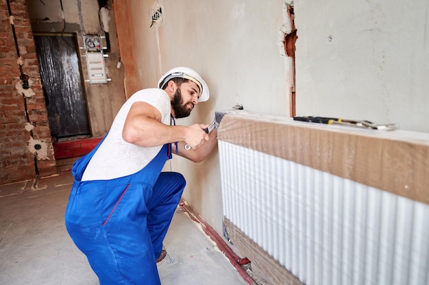 Giovane idraulico che indossa una tuta blu con casco bianco che installa il radiatore dell'acqua lavorando con la chiave