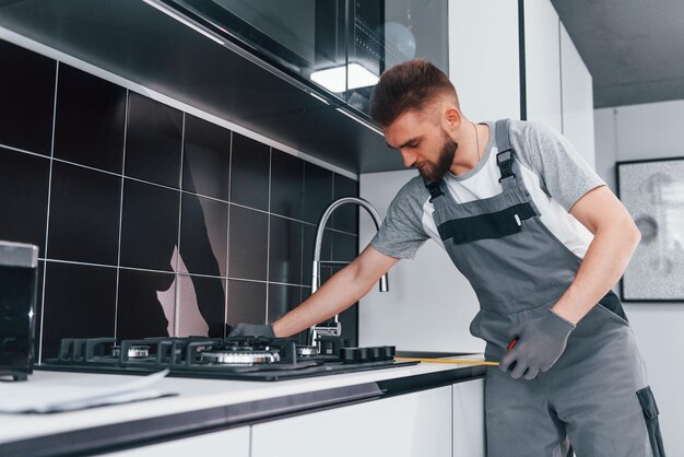 Giovane idraulico professionista in grigio tavolo di misurazione uniforme utilizzando il metro in cucina