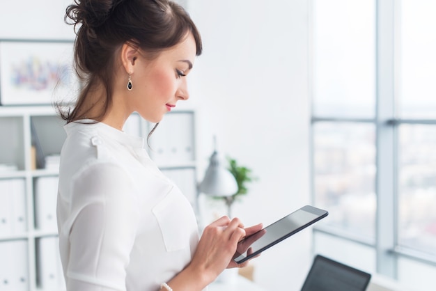 Giovane impiegato femminile sorridente al suo posto di lavoro leggendo, passando in rassegna i messaggi di annunci di notizie utilizzando il computer tablet durante una pausa.