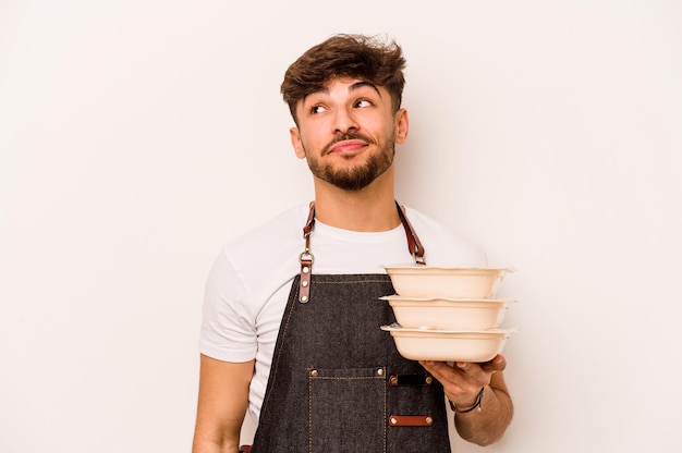 Giovane impiegato ispanico che tiene un tupperware isolato su sfondo bianco sognando di raggiungere obiettivi e scopi