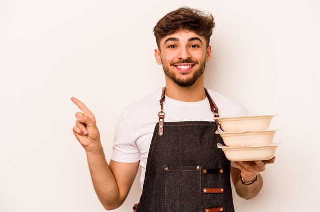 Giovane impiegato ispanico che tiene un tupperware isolato su sfondo bianco sorridente e rivolto da parte mostrando qualcosa in uno spazio vuoto