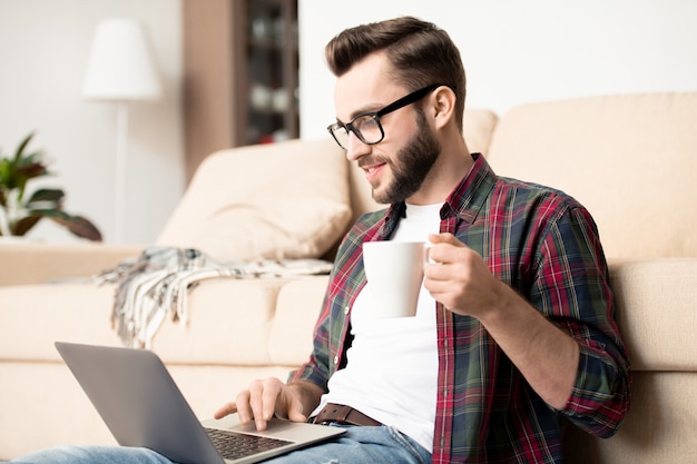 Giovane impiegato occasionale con la tazza di tè e il computer portatile che si siede dallo strato nel soggiorno e che lavora a distanza