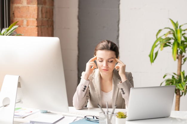 Giovane impiegato stanco in abbigliamento formale che tocca le sue tempie mentre cercava di concentrarsi durante il lavoro al computer in ufficio