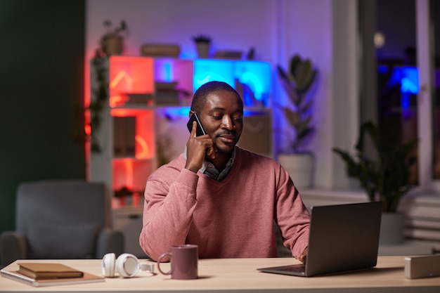 Giovane imprenditore africano seduto al suo posto di lavoro digitando sul laptop e ha una conversazione al telefono lavorando fino a sera in ufficio