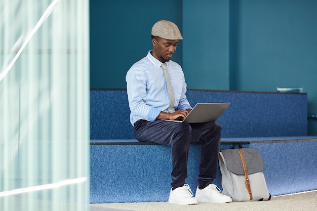 Giovane imprenditore africano seduto in corridoio e digitando sul computer portatile in ufficio
