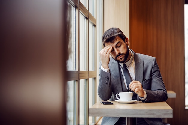 Giovane imprenditore barbuto seduto nella caffetteria accanto alla finestra e avendo mal di testa. Pensa che il caffè del mattino potrebbe aiutare.