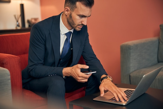 Giovane imprenditore calmo e concentrato in abito seduto con uno smartphone e guardando lo schermo di un laptop sul tavolino da caffè durante la digitazione