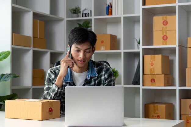 Giovane imprenditore che conduce una telefonata d'affari mentre gestisce le vendite online utilizzando un portatile in uno spazio di lavoro moderno con scatole di prodotti
