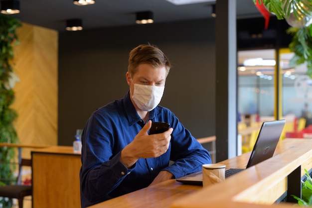 Giovane imprenditore con maschera utilizzando il telefono e seduto con la distanza all'interno della caffetteria