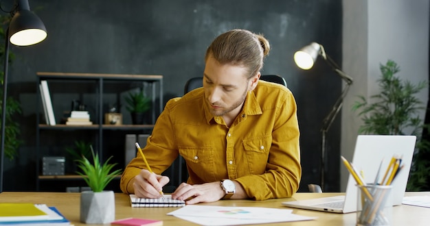 Giovane imprenditore, seduto al tavolo in ufficio e scrivere nelle note in taccuino, lavorando su un computer portatile.