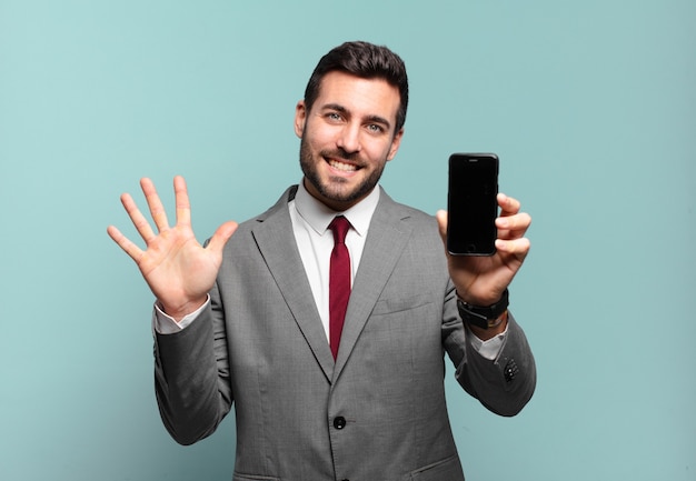 Giovane imprenditore sorridente e guardando amichevole, mostrando il numero cinque o quinto con la mano in avanti, il conto alla rovescia e mostrando il suo schermo del telefono