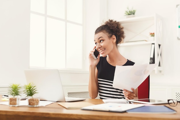 Giovane imprenditrice afro-americana sorridente che parla al telefono con dei documenti, seduta al moderno posto di lavoro dell'ufficio. Consulenza aziendale, copia spazio